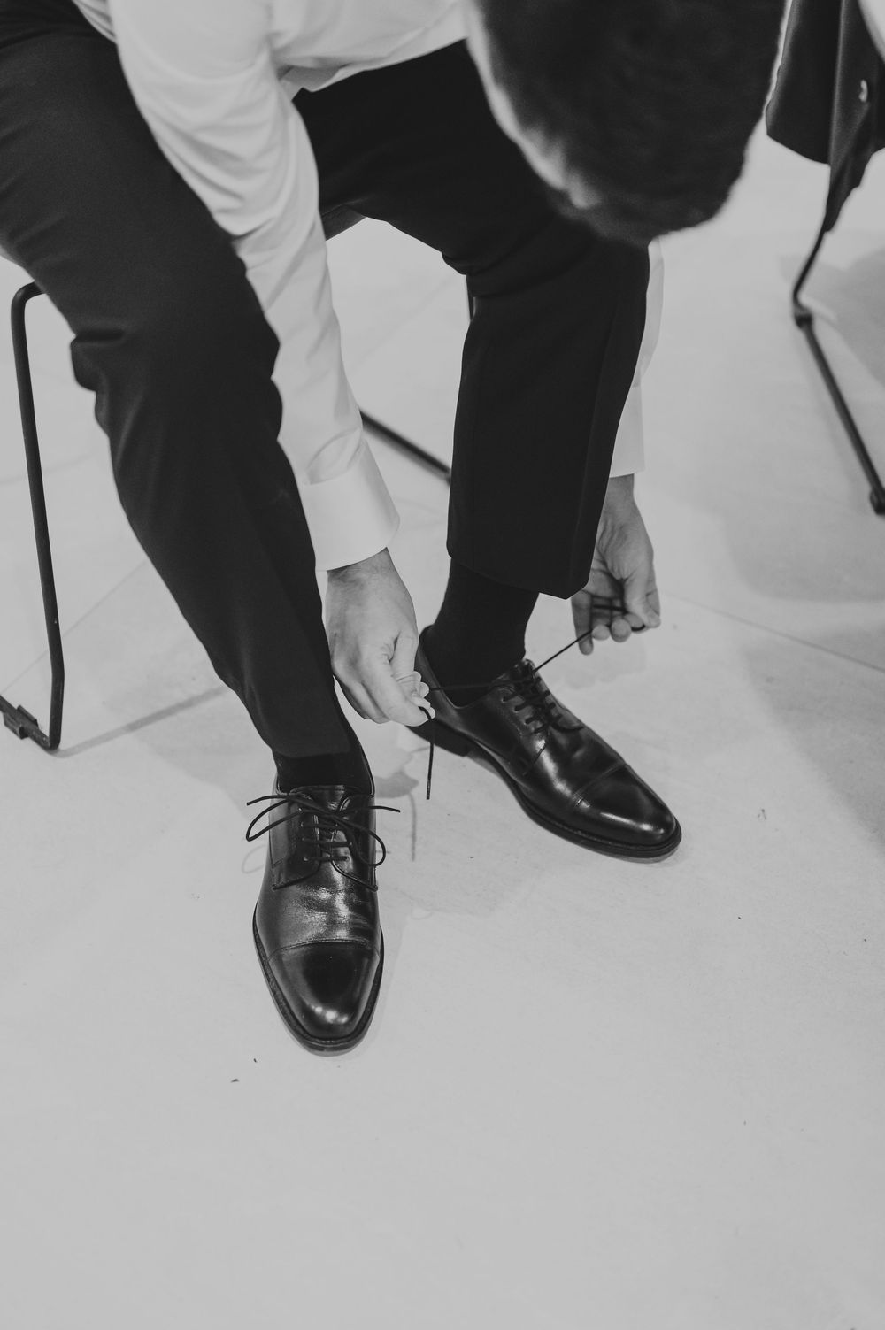 Black and white photo of groom tying laces on classic black Oxford dress shoes.