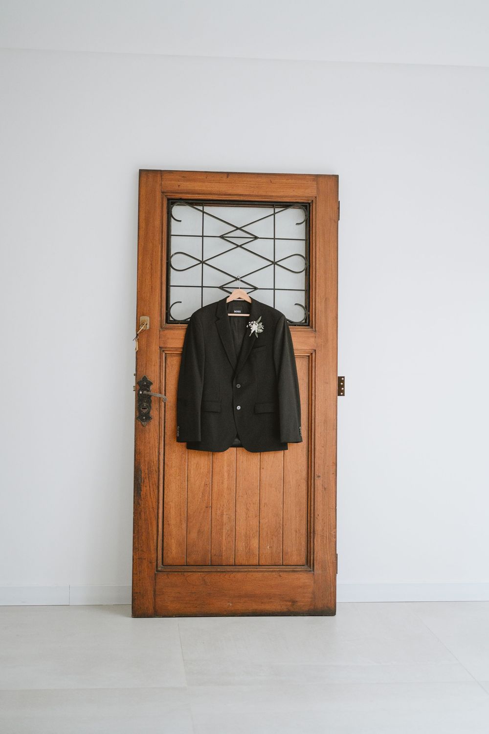 Dark suit jacket with floral boutonniere on wooden hanger displayed against rustic antique wooden door.