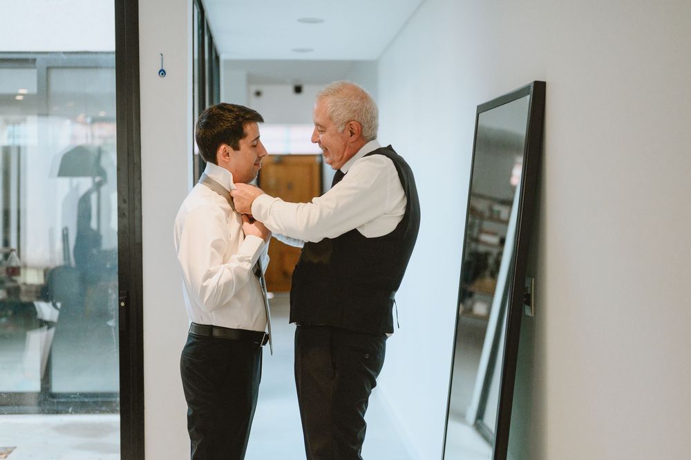 Older man helping young groom adjust tie during wedding preparations in modern hallway.