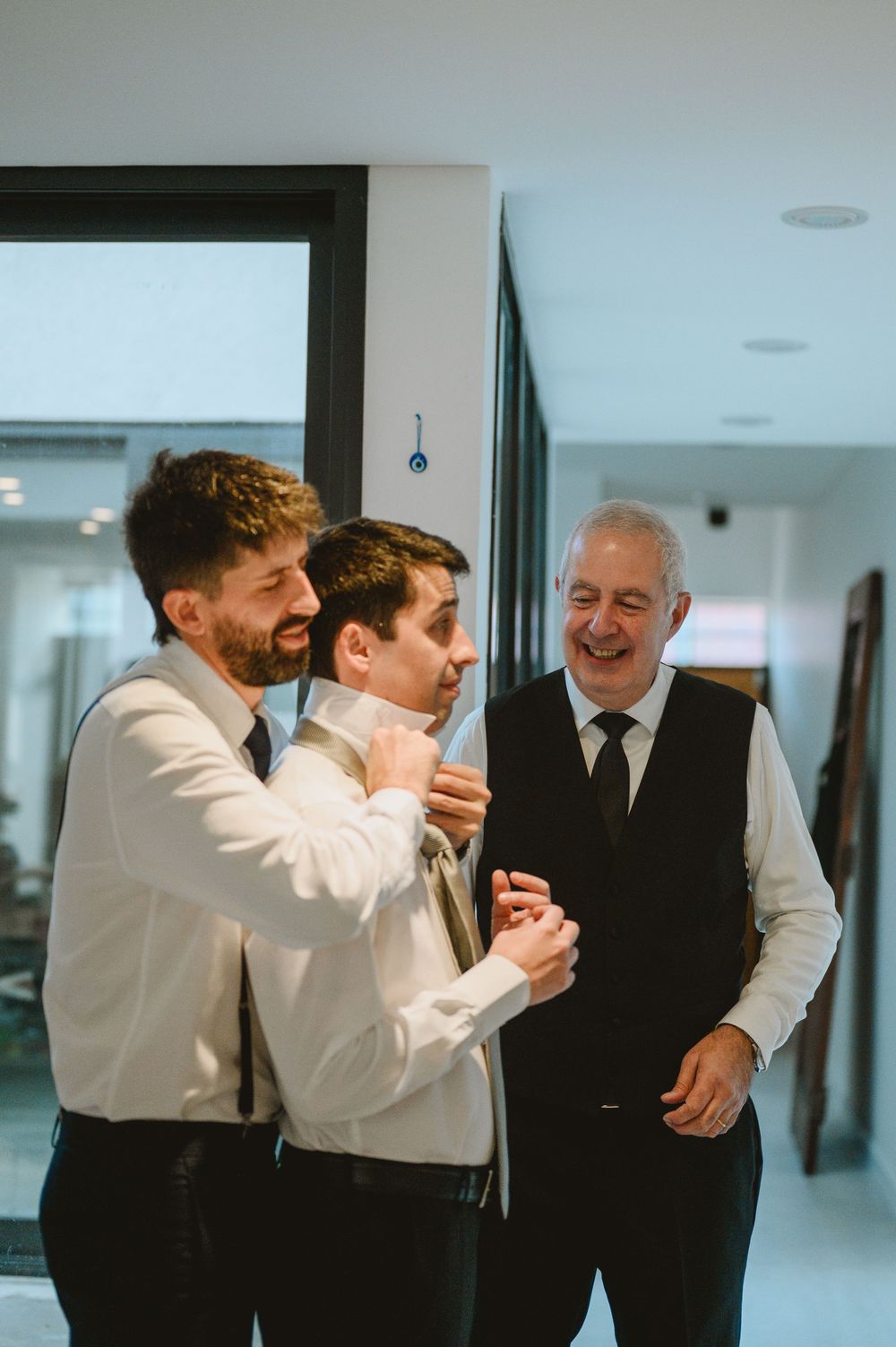 Groom laughing with groomsman and father while getting tie adjusted before wedding ceremony.