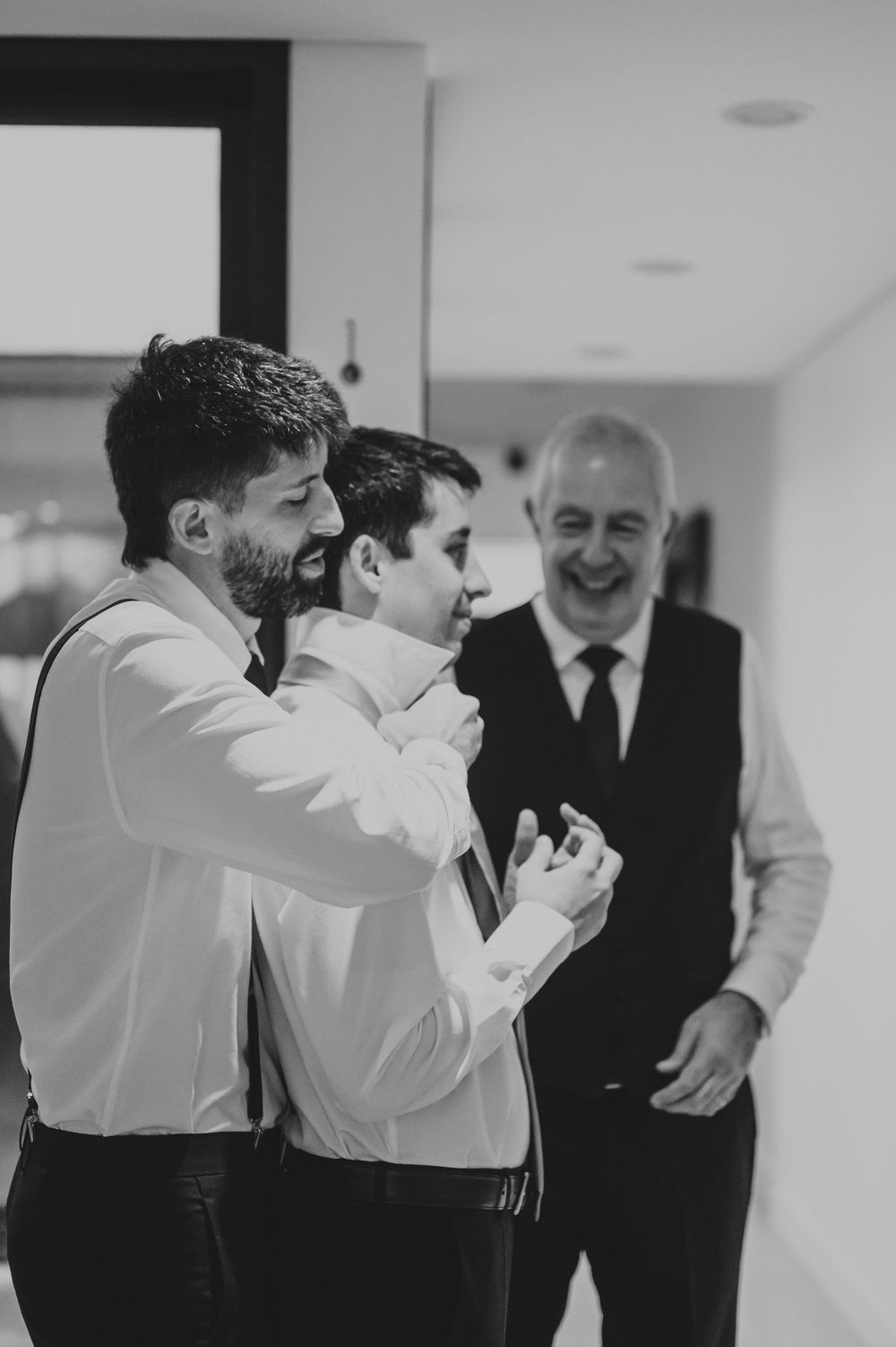 Black and white photo of groomsmen laughing while helping groom fix tie before wedding.