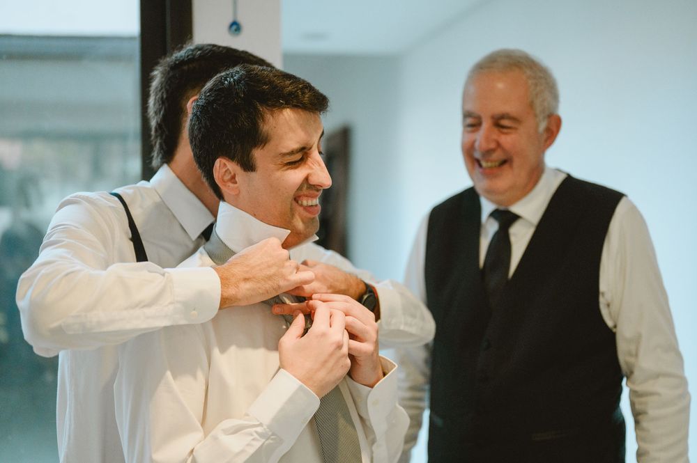 Groom smiling while groomsman helps pin boutonniere as father watches during wedding preparations.