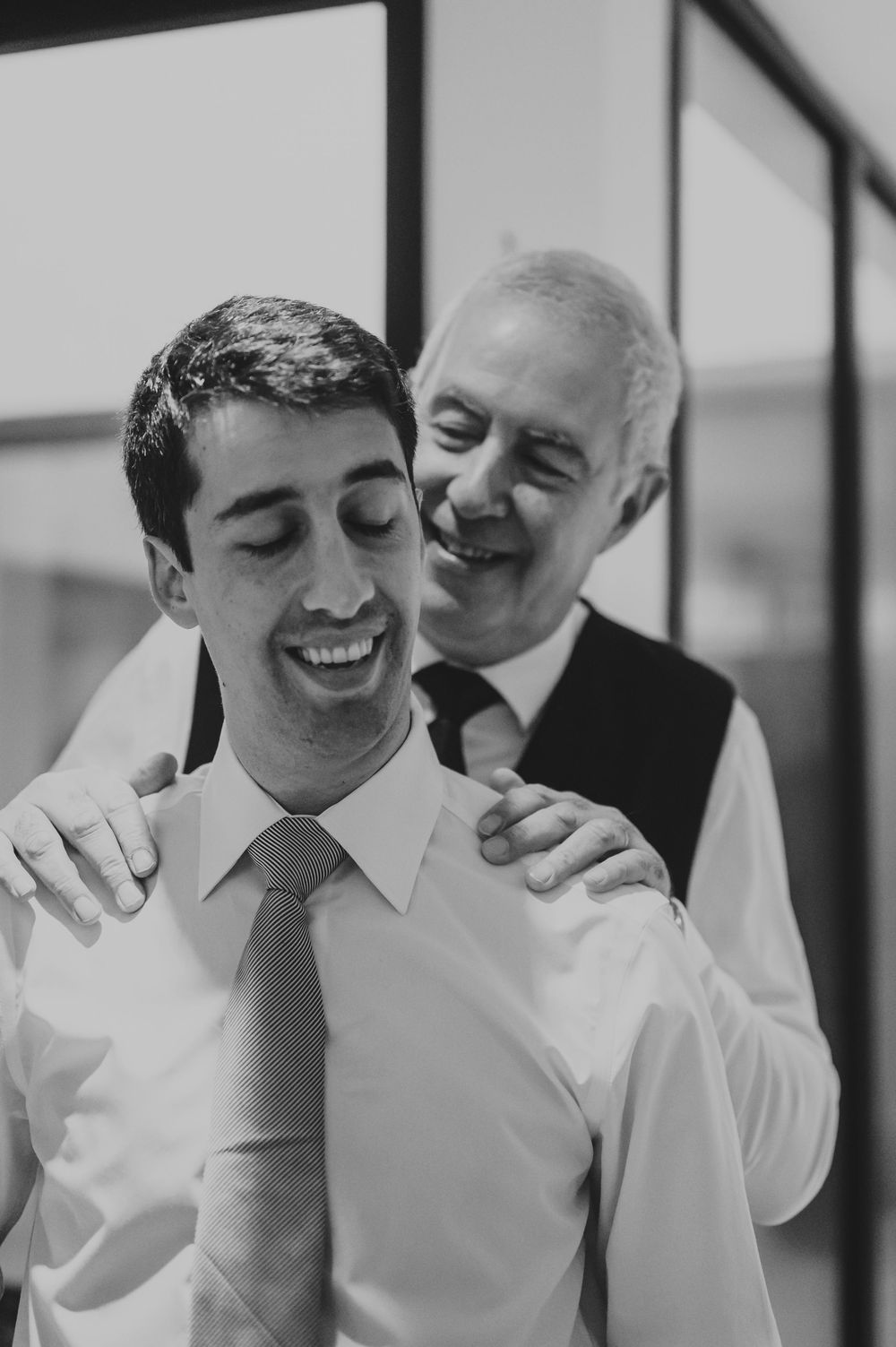Black and white portrait of smiling groom with father embracing his shoulders before wedding.