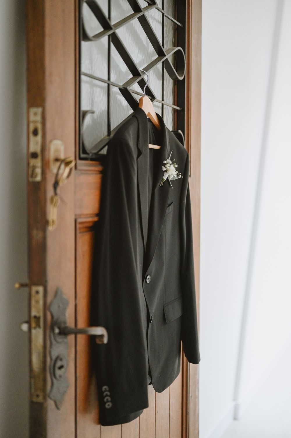 Close-up of dark groom suit jacket with boutonniere hanging on ornate vintage wooden door.