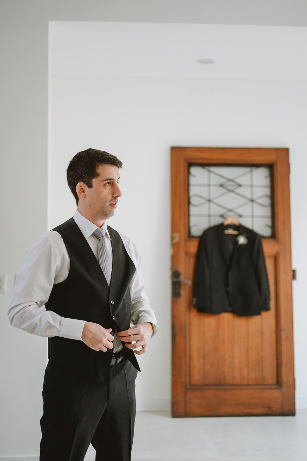 Groom in white shirt and black vest buttoning up while his suit jacket hangs on a rustic wooden door behind him.
