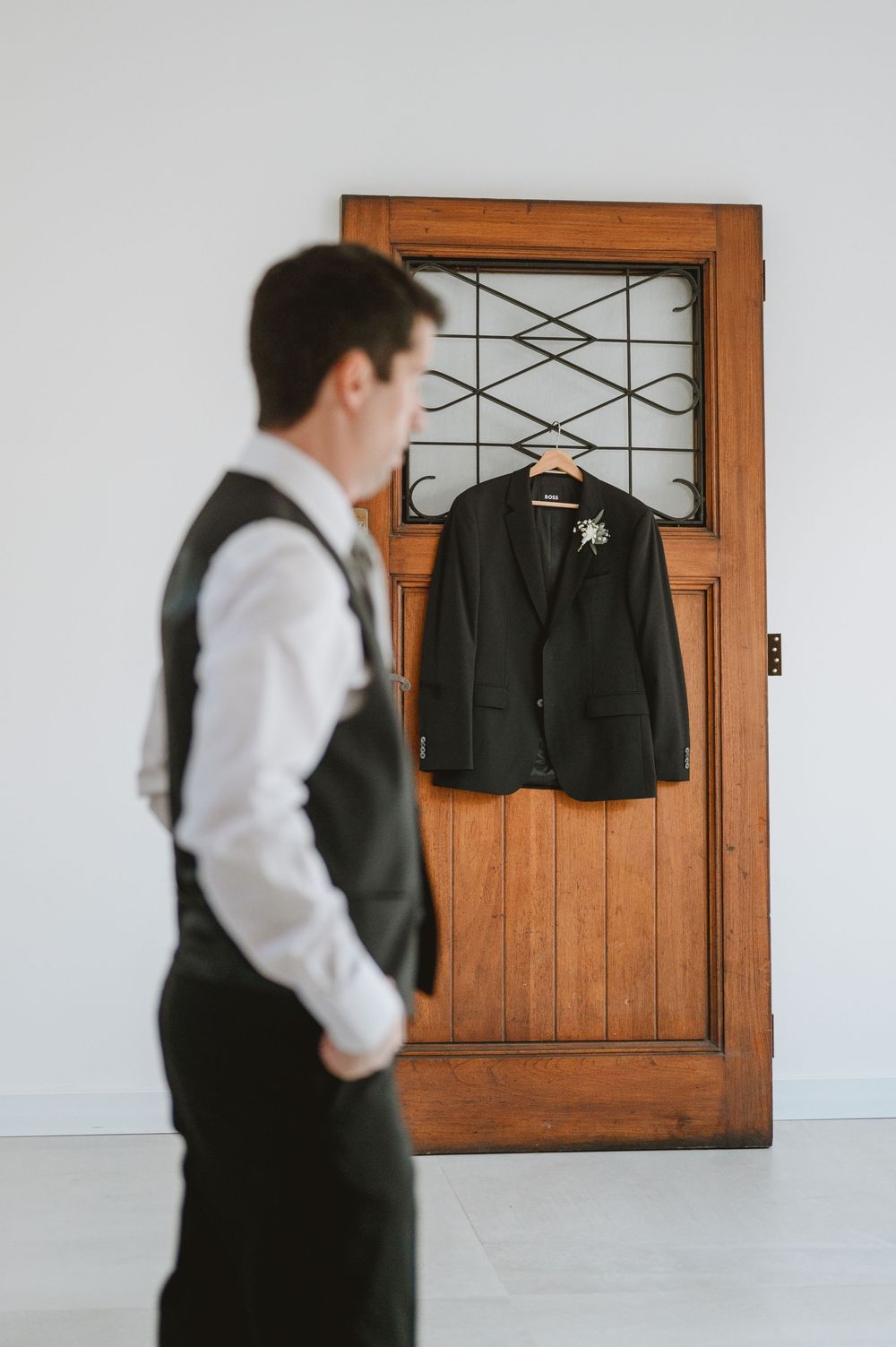 Groom in black vest and white shirt gazes at his suit jacket with boutonniere hanging on a vintage wooden door.