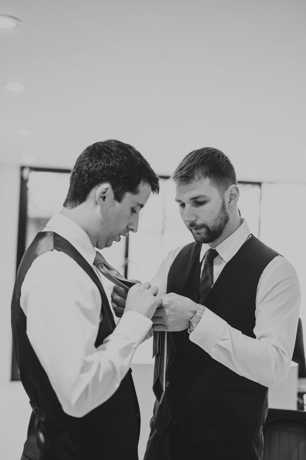 Two groomsmen in black vests help each other straighten their ties in a black and white wedding preparation photo.