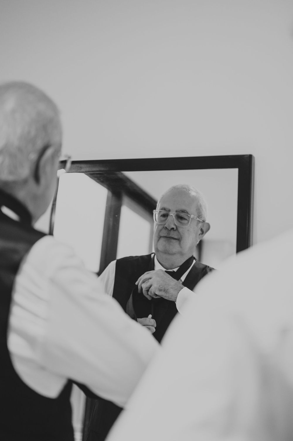 Elderly man in black vest adjusts his bow tie while looking at his reflection in a mirror before a wedding.