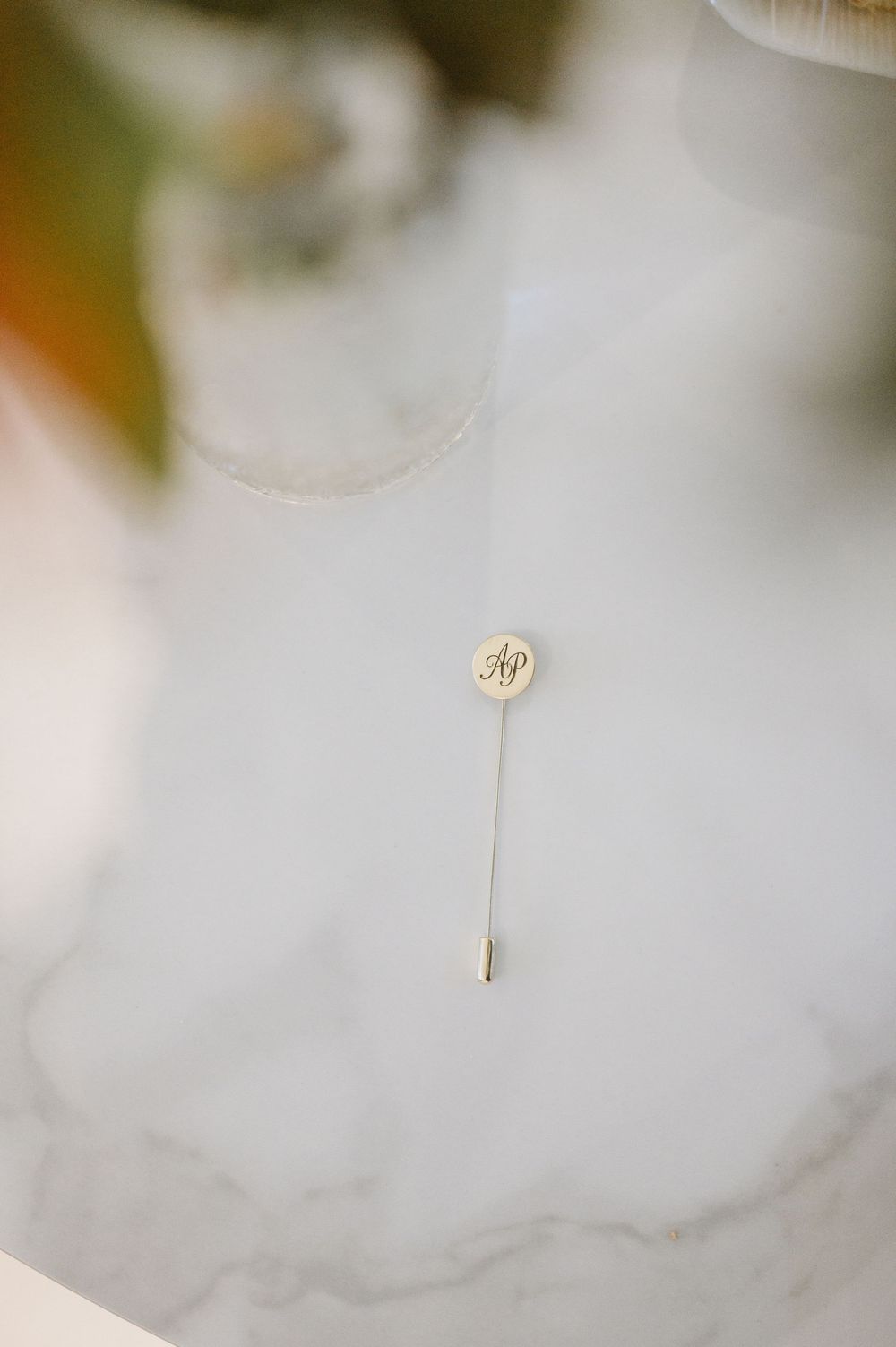 Gold monogrammed lapel pin on stick displayed on marble surface for groom wedding details.
