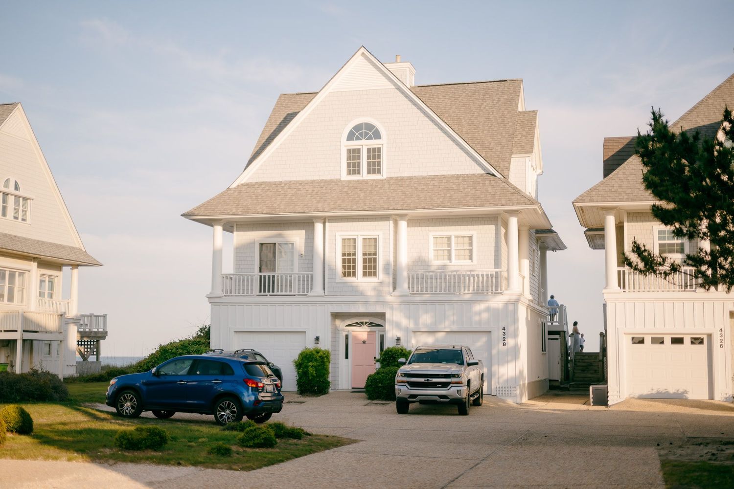 White coastal beach house with blue SUV and silver truck parked in driveway on a sunny day.