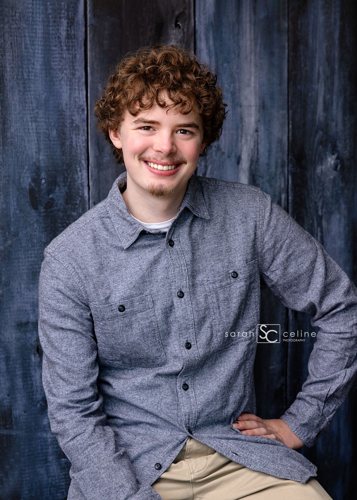 Smiling young man with curly hair wearing a gray button-up shirt, posing against a rustic blue wooden backdrop.