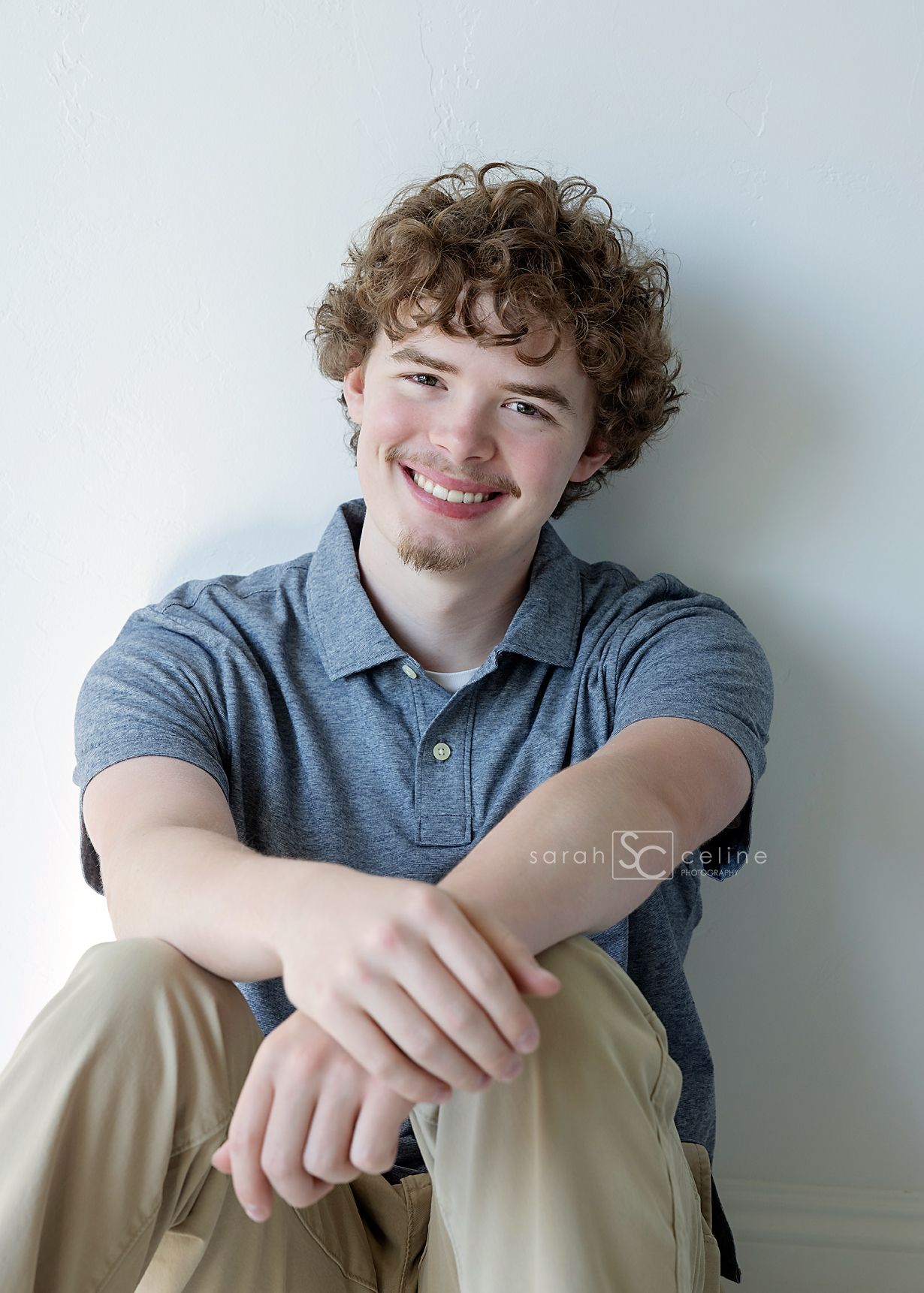 Smiling young man with curly hair wearing a gray polo shirt and khaki pants, sitting casually against a white wall.