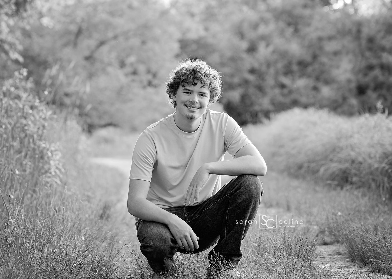 A smiling teenage boy crouches on a dirt path surrounded by tall grass and trees in a black and white outdoor portrait.