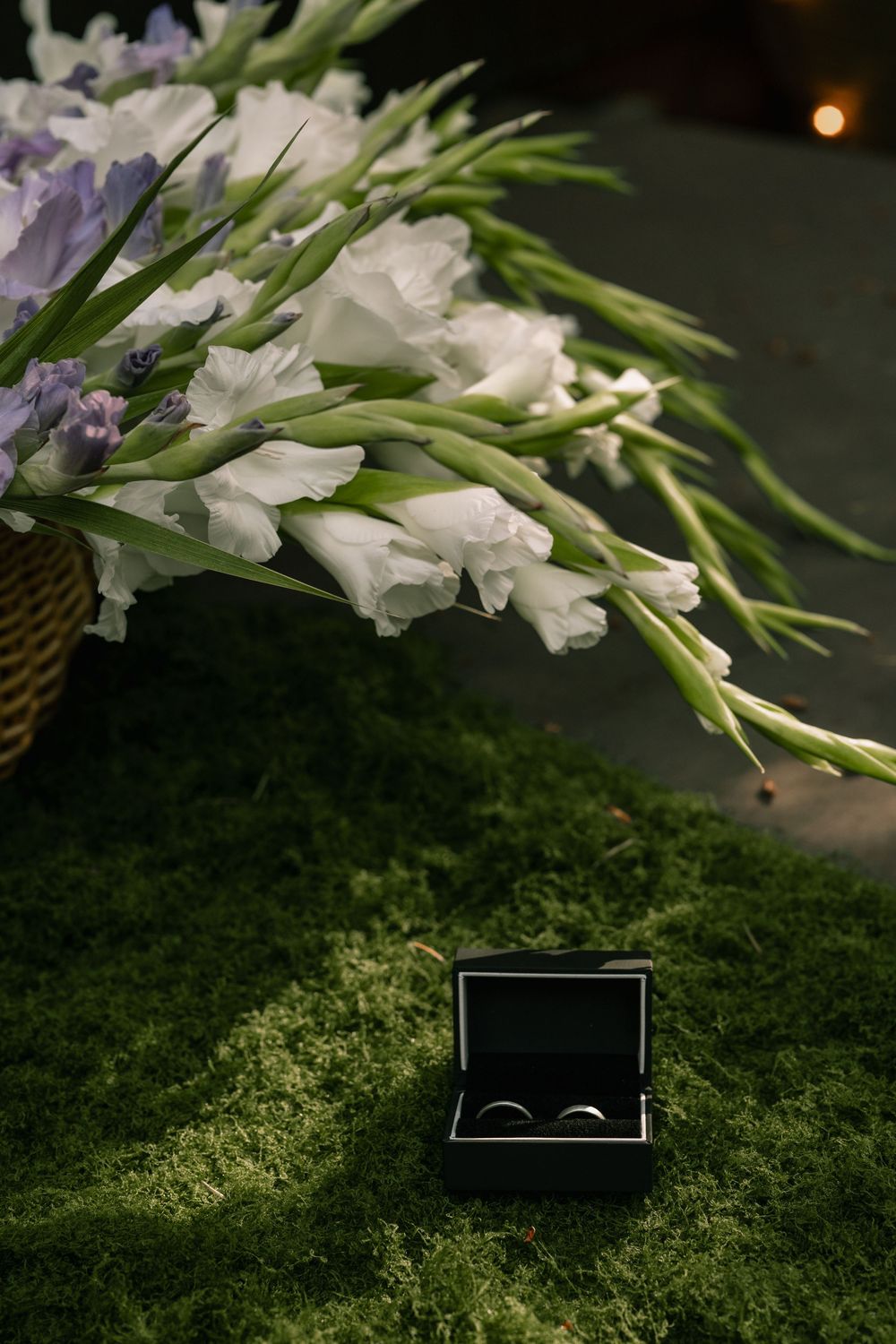 Wedding rings in black box on green moss beside white gladiolus bouquet in wicker basket.