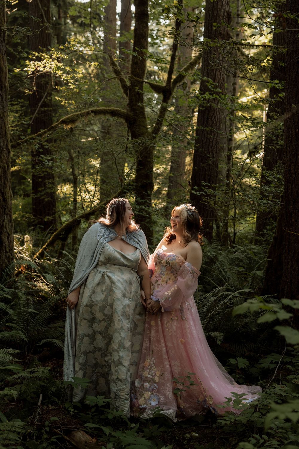 Two brides in elegant gowns share a romantic moment in a lush forest setting.