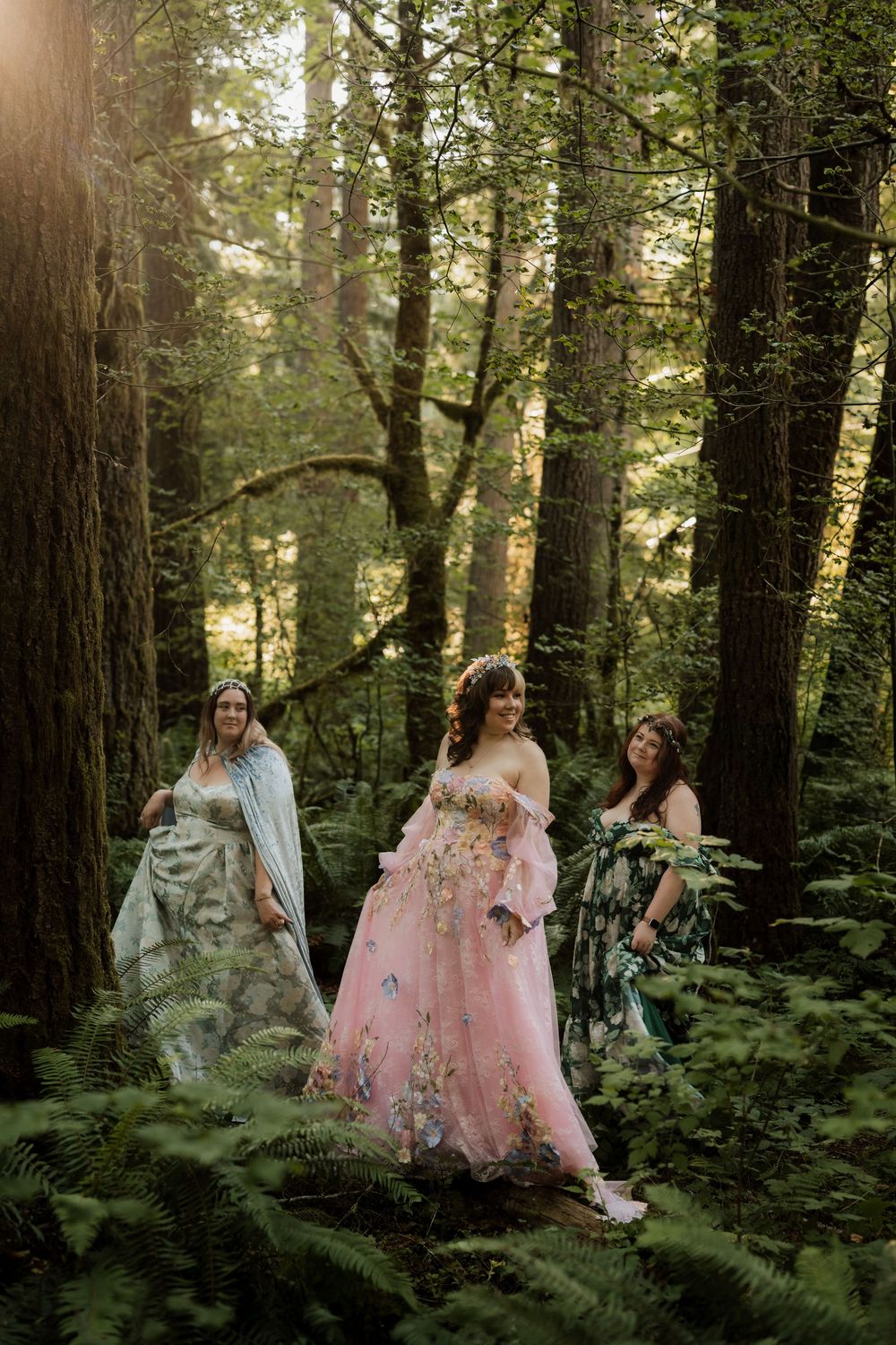 Three women in floral gowns walk through a sunlit forest, surrounded by ferns and tall trees.