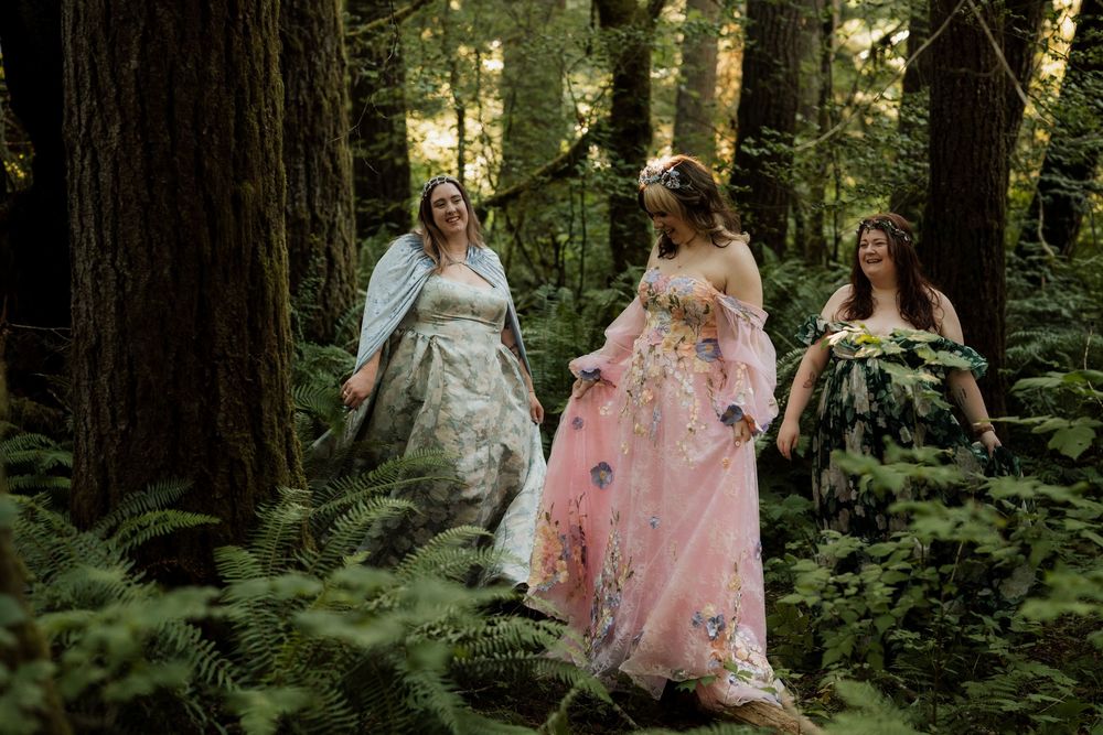 Three women in floral fantasy gowns walk through a lush forest surrounded by ferns and towering redwood trees.