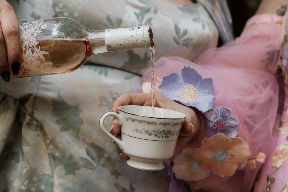Rose wine being poured into a vintage floral teacup held by a woman in a floral dress.