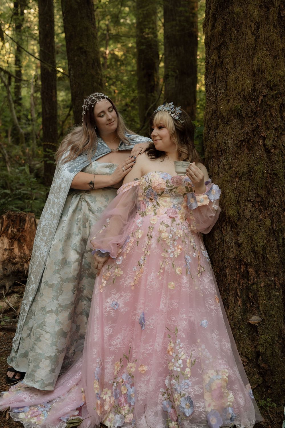 Two women in elegant fantasy-inspired gowns and floral crowns posing in a lush forest setting.