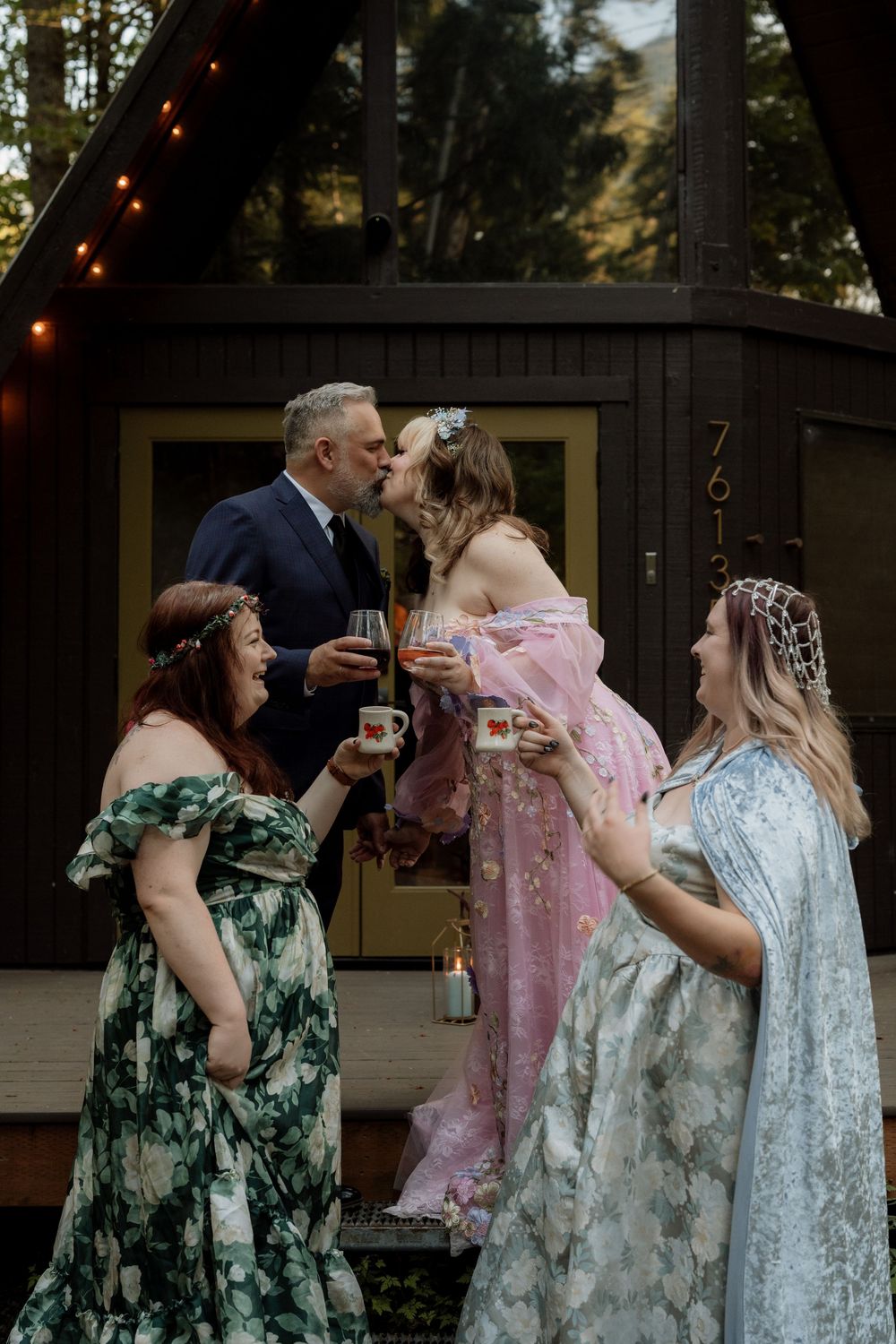 Couple kisses at rustic A-frame cabin while two women in floral gowns toast with drinks at outdoor wedding celebration.