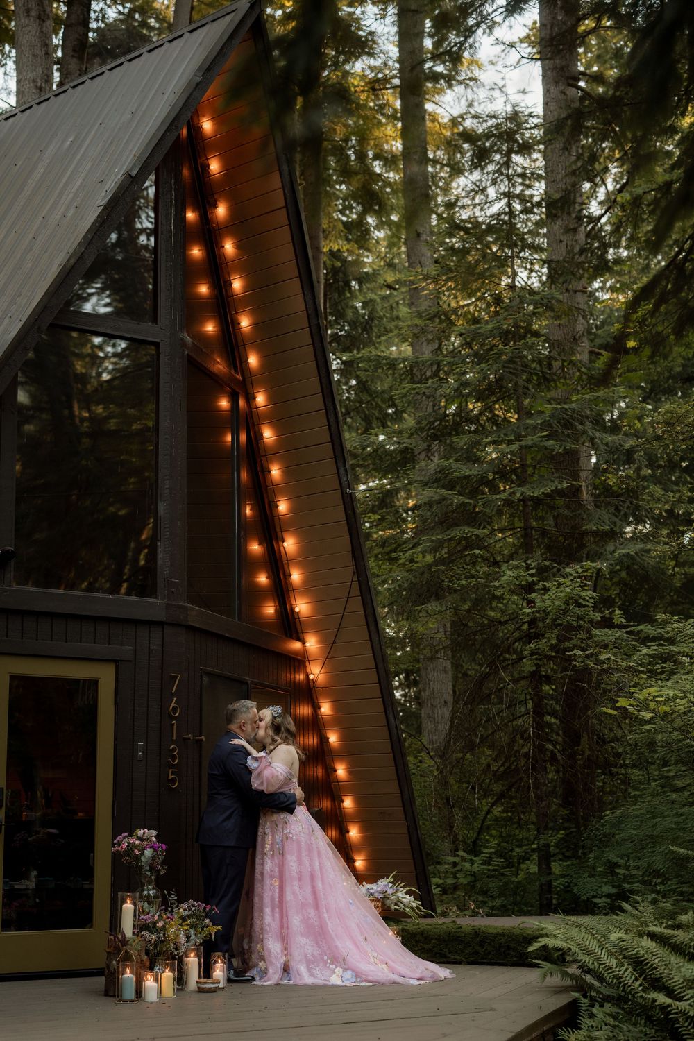 Couple embraces on the porch of an A-frame cabin adorned with warm string lights.