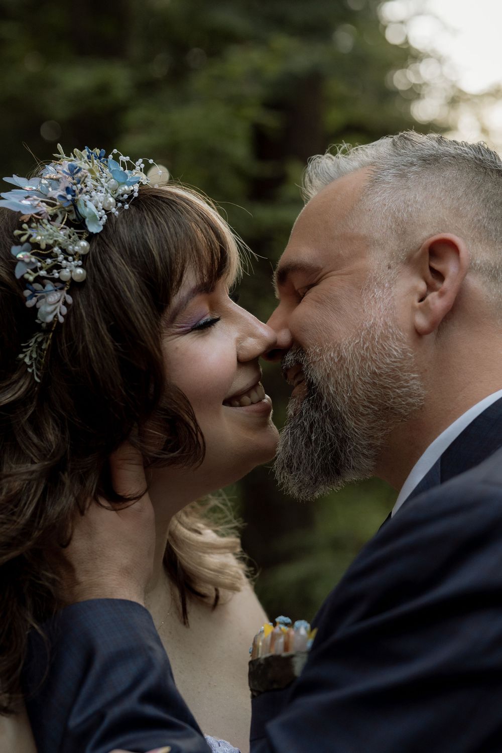 Bride with floral crown smiles as groom leans in for a kiss outdoors.