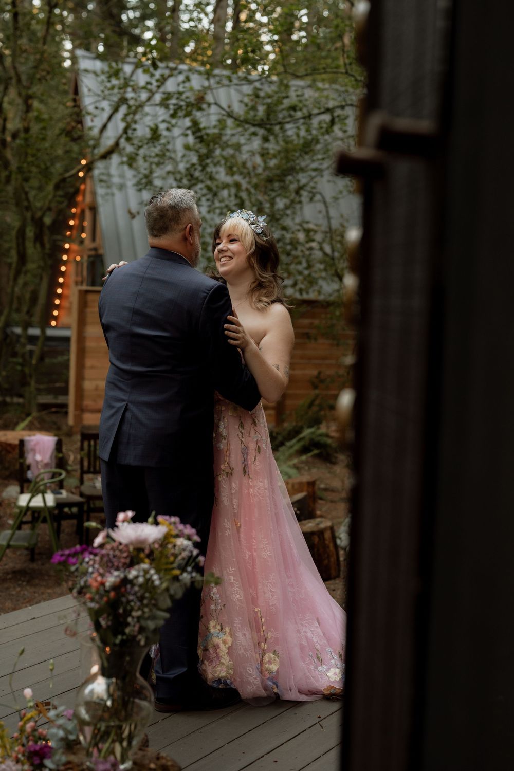 Bride in pink floral gown smiles at partner in navy suit while dancing outdoors at a romantic garden wedding.