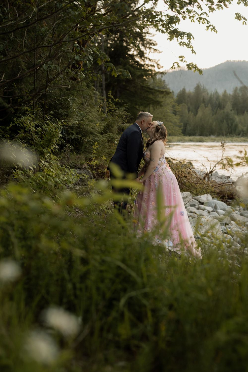 Couple sharing a romantic kiss in a pink gown by a misty river, surrounded by lush green trees and mountains at dusk.