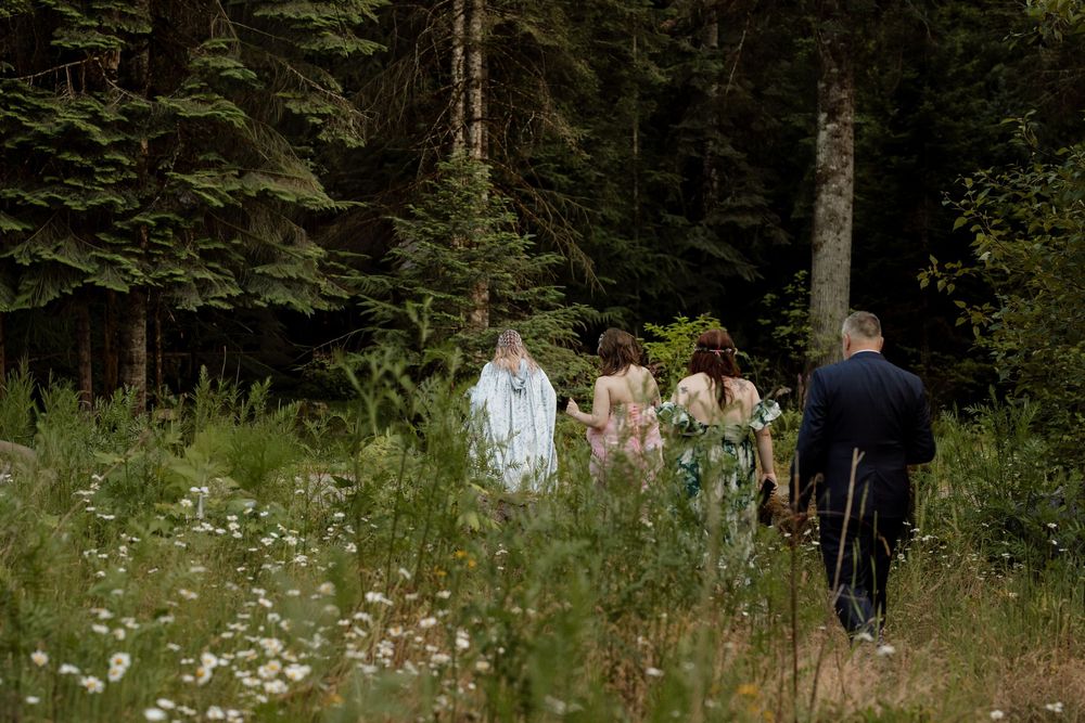Wedding party walking into a lush forest clearing surrounded by wildflowers and tall evergreen trees.