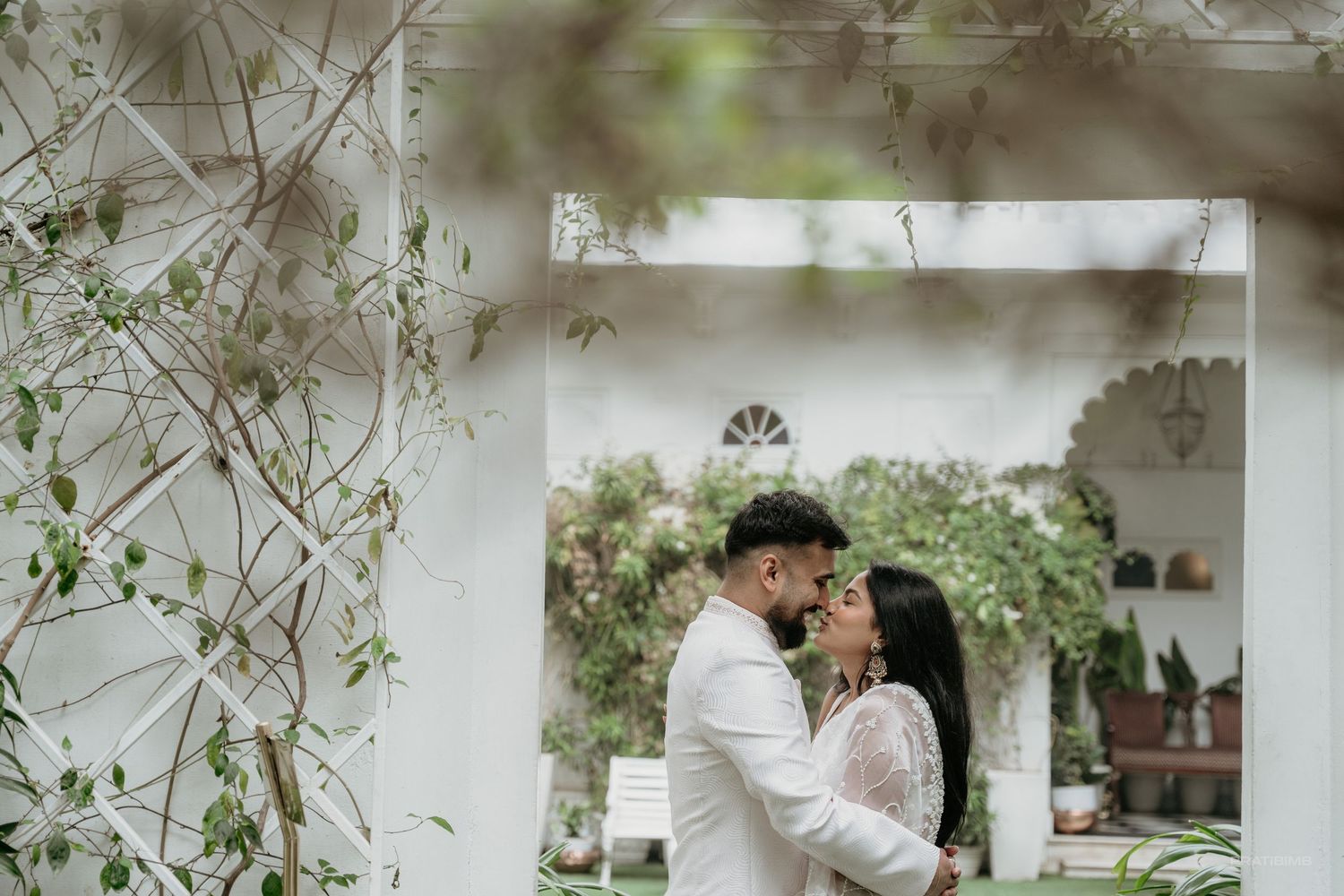 Couple sharing a romantic moment in a lush garden setting, surrounded by greenery and white architecture.