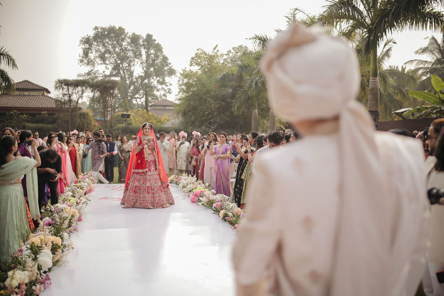 Bride in red lehenga walks down floral aisle at outdoor Indian wedding ceremony with groom waiting ahead.