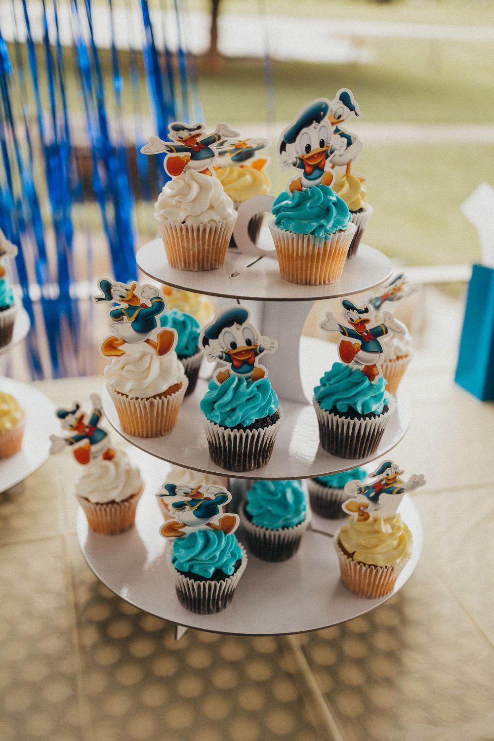 Three-tier cupcake stand with Donald Duck toppers, blue and white frosted cupcakes at a birthday party.