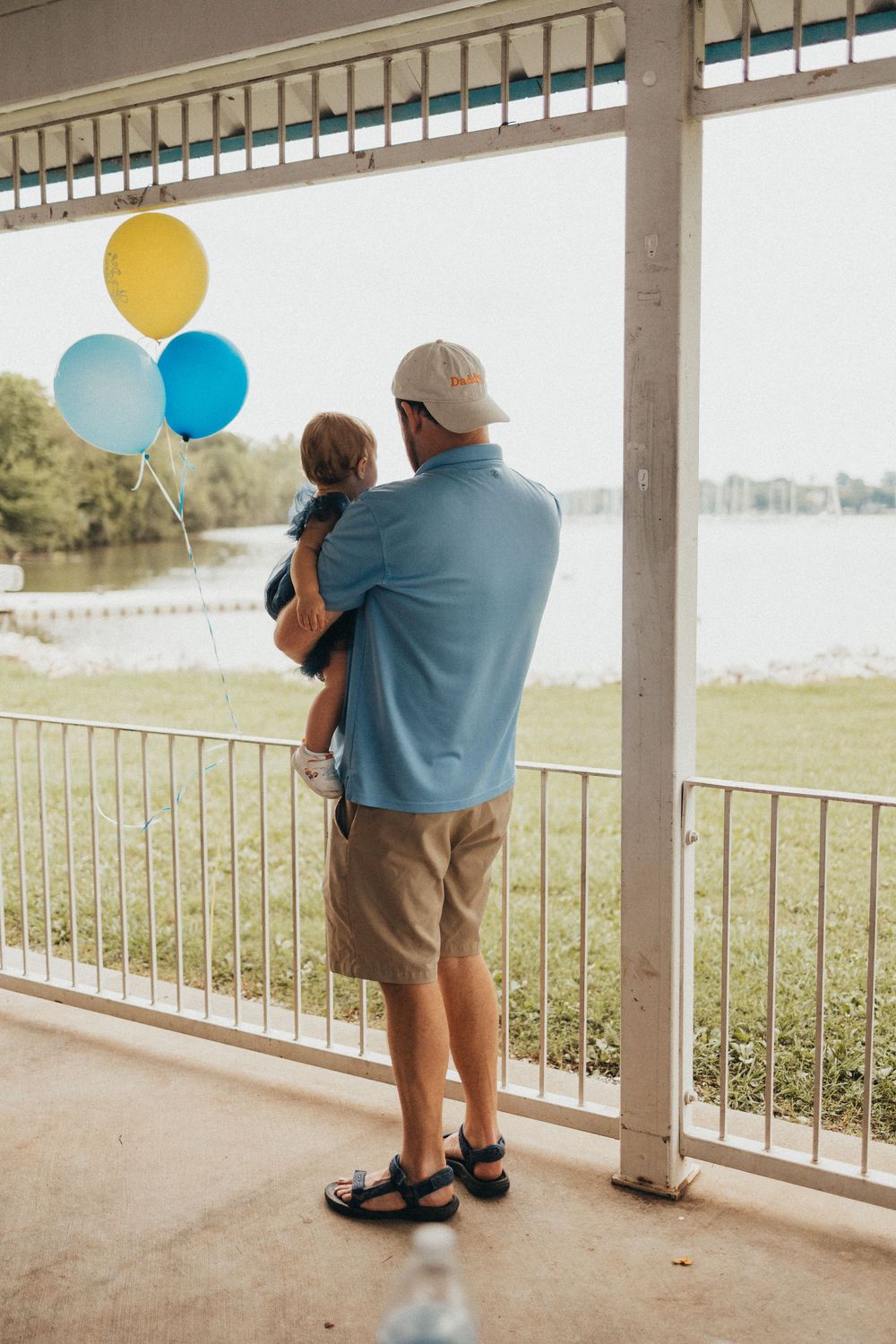 Man holding a child with blue and yellow balloons on a porch overlooking a serene waterfront.