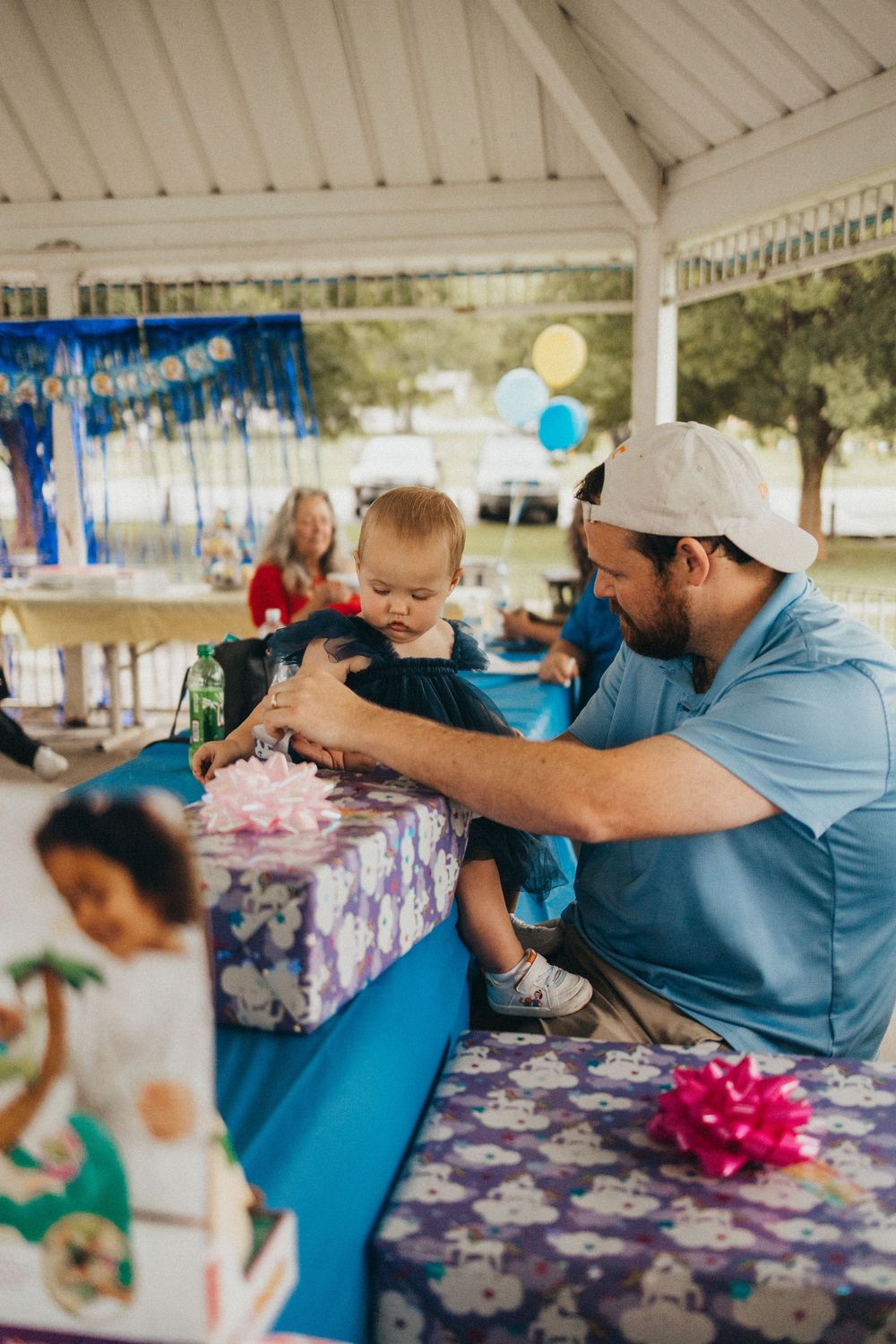 A man in a blue shirt helps a baby open a wrapped gift at an outdoor birthday party under a pavilion.
