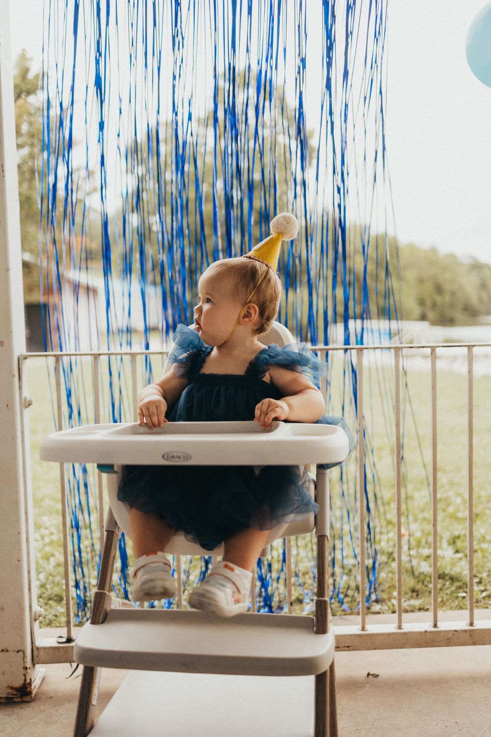 Baby girl in blue tutu dress and gold party hat sits in high chair with blue fringe backdrop for first birthday.