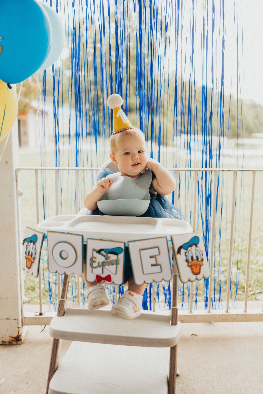 Baby in birthday hat sits in high chair with ONE banner and blue streamers at a Donald Duck first birthday party.