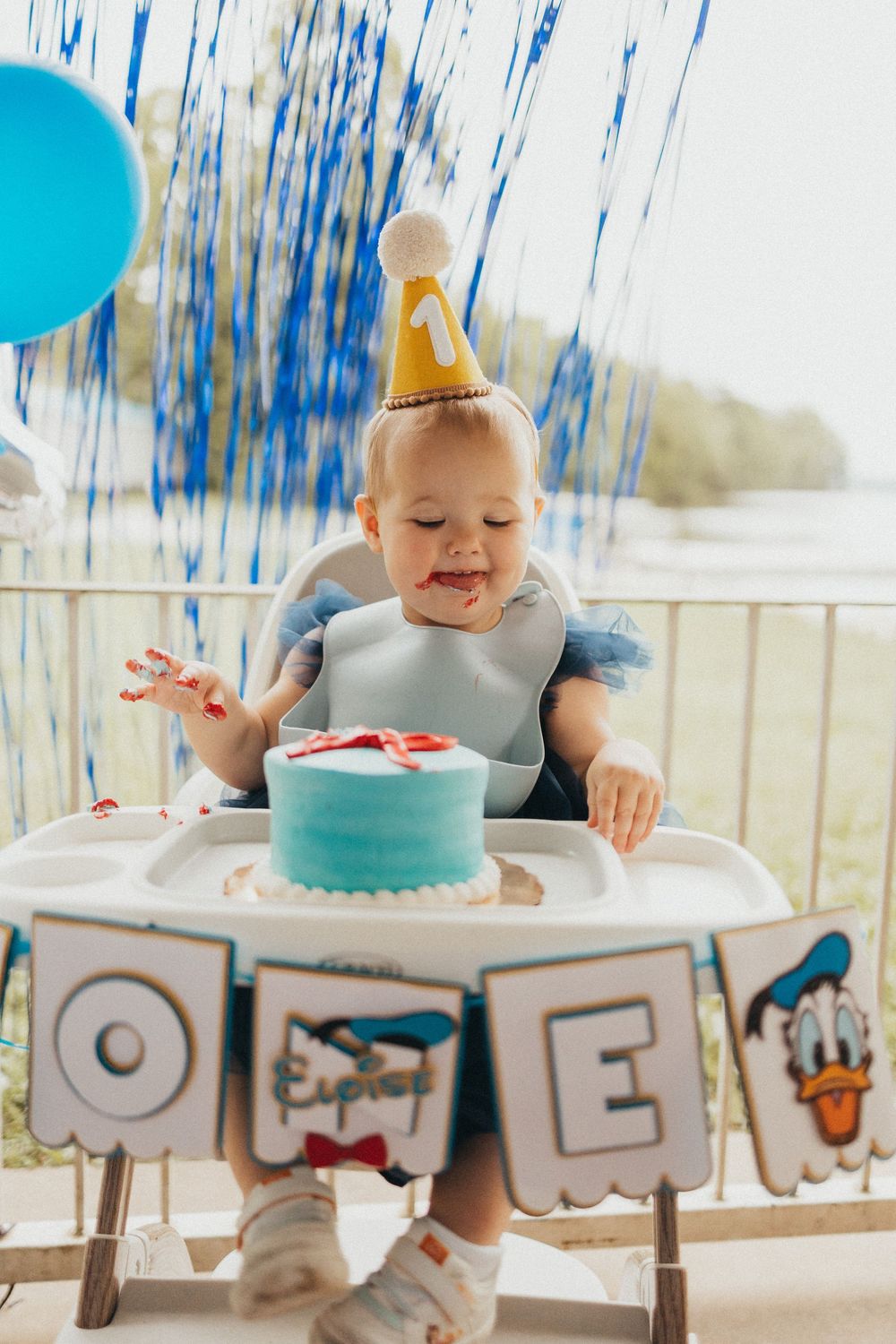 Baby in party hat smashes first birthday cake in high chair, decorated with blue streamers and ONE banner.
