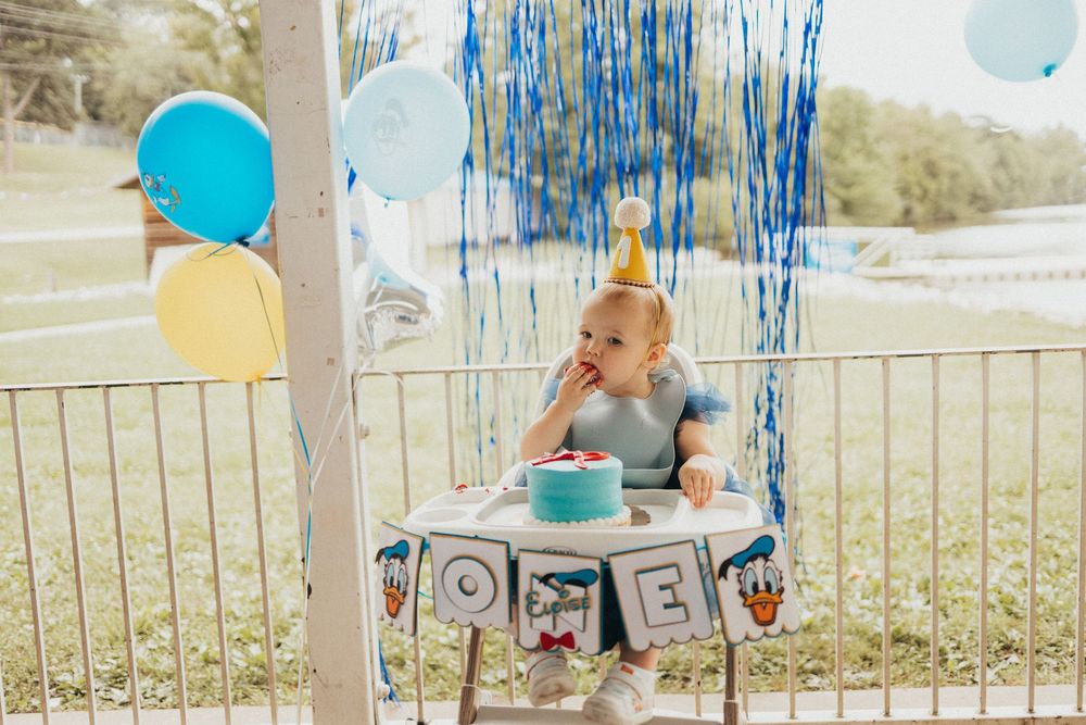 Baby in gold party hat enjoys first birthday cake smash with blue balloons and ONE letter blocks outdoors.