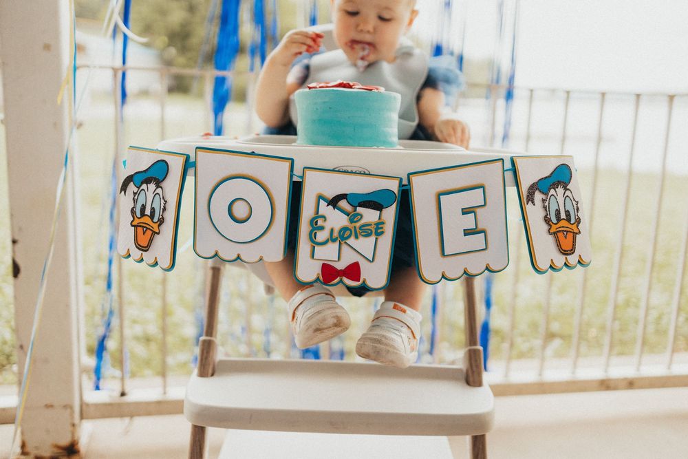 Baby in high chair celebrating first birthday with Donald Duck ONE banner and blue smash cake.