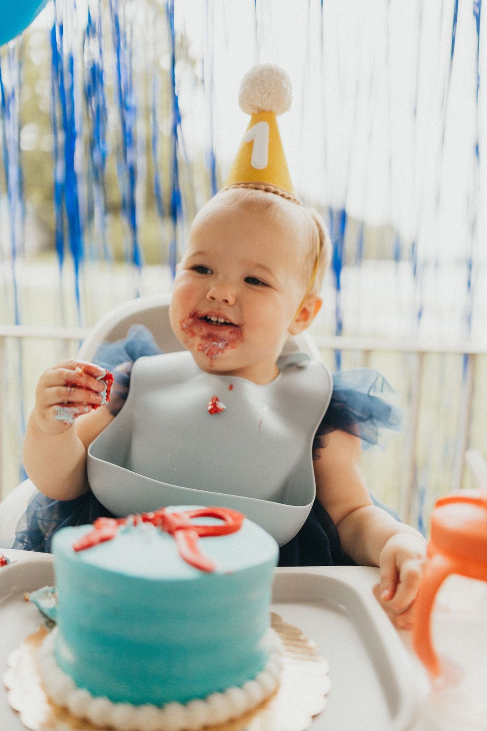 A happy baby wearing a party hat celebrates their first birthday with a blue smash cake, face covered in frosting.