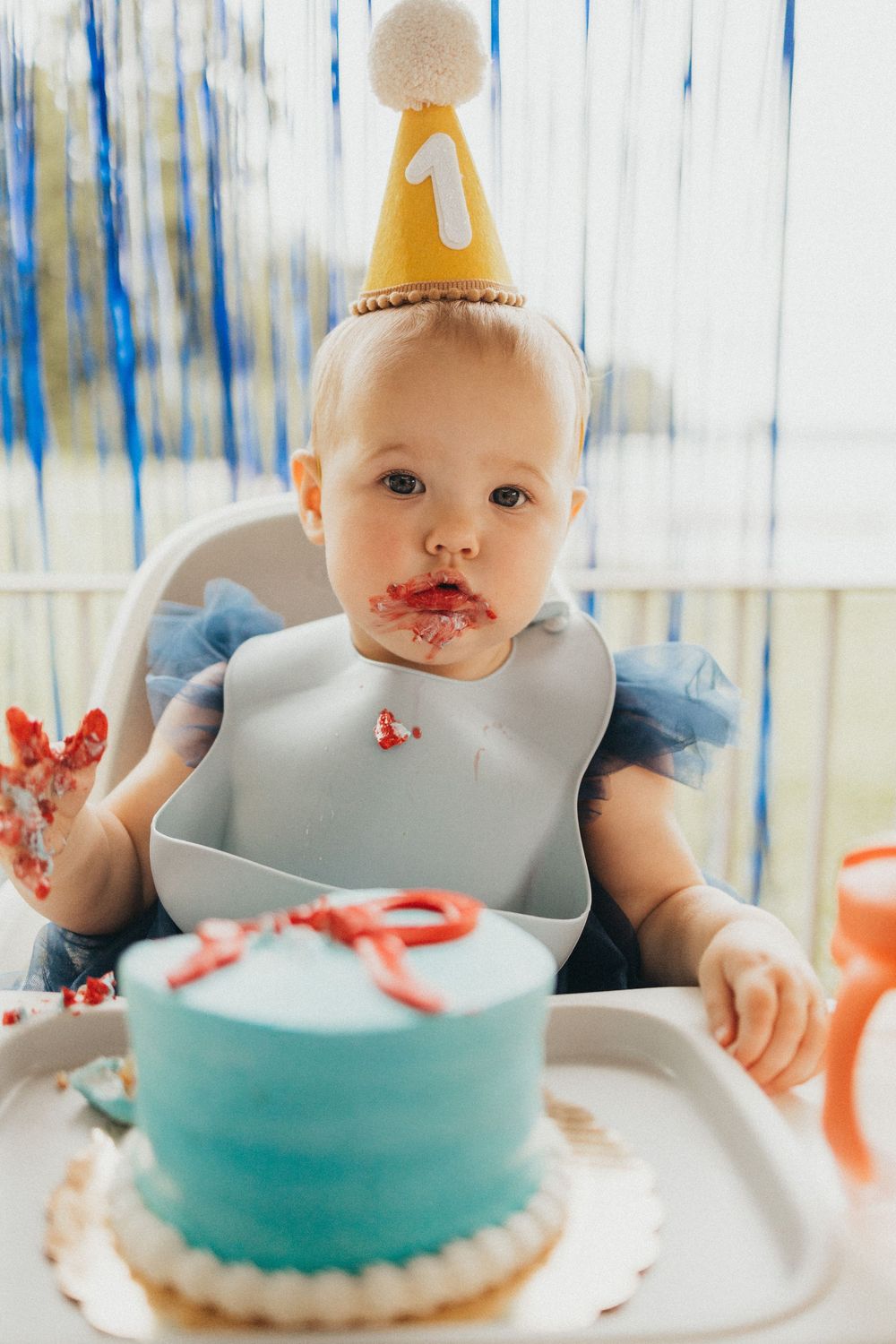 Baby in a number 1 party hat with cake smash frosting on face sits in high chair with blue birthday cake.