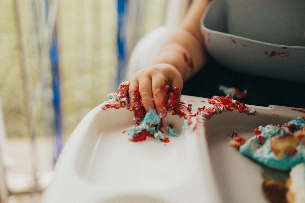 Baby's hand covered in red and blue cake smash frosting on white high chair tray during first birthday.
