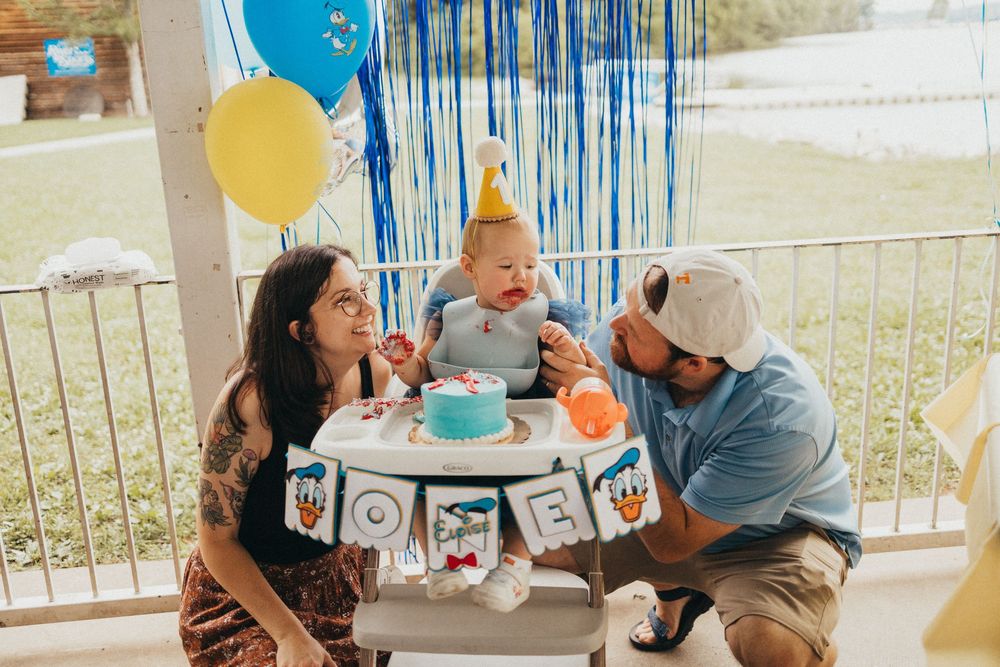 Family celebrating baby's first birthday with cake smash, blue balloons, and party decorations outdoors.