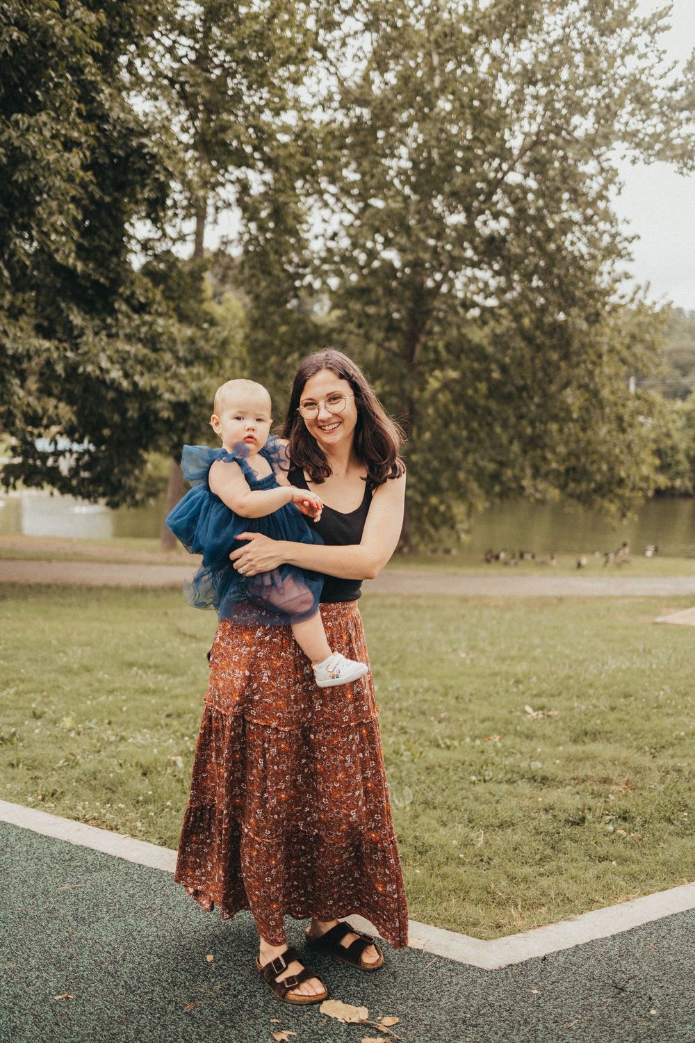A smiling woman in a floral skirt holds a baby in a blue dress outdoors in a green park setting.