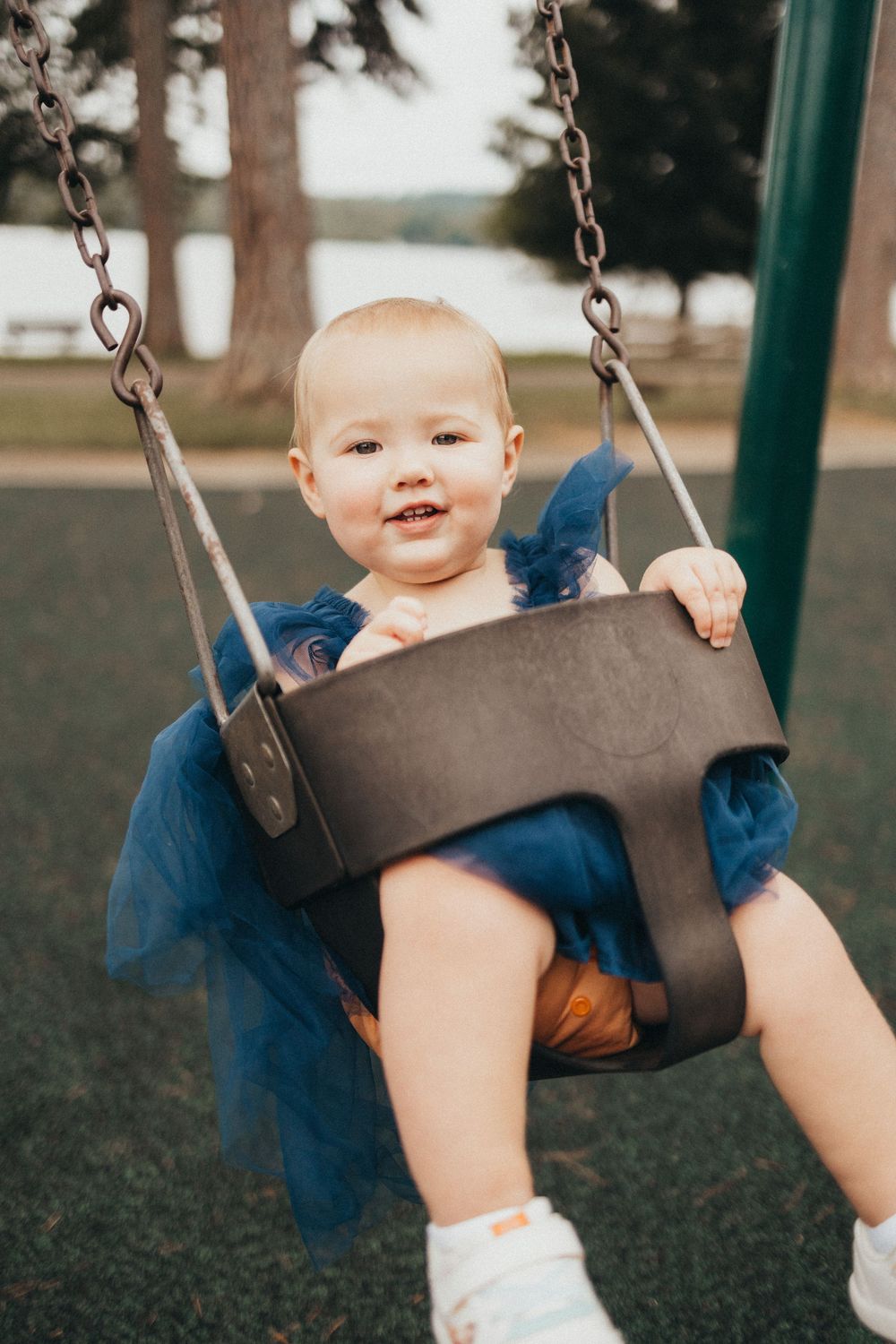 A happy baby in a blue tulle dress smiles while sitting in a toddler bucket swing at a park playground.