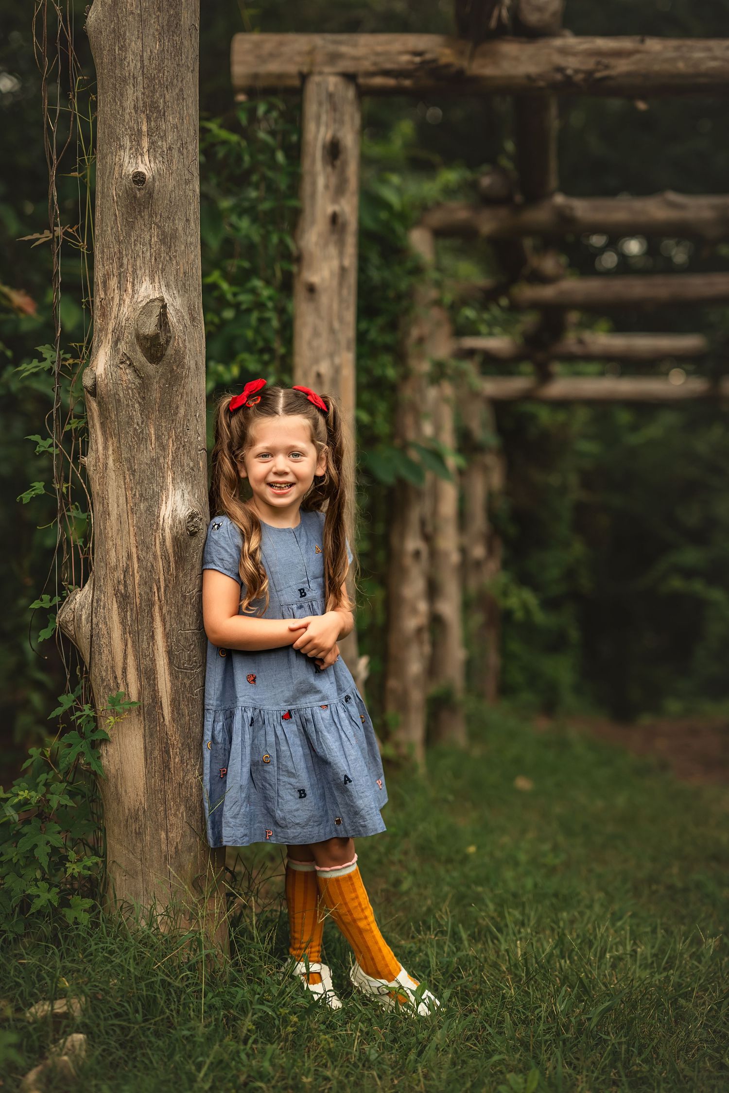 Young girl in blue dress and yellow socks leaning against rustic wooden post in a lush green garden.