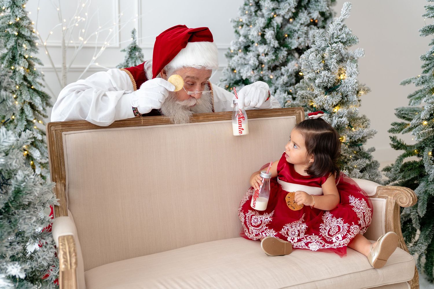 A toddler girl in a red dress sits on a beige sofa with Santa Claus beside decorated Christmas trees.