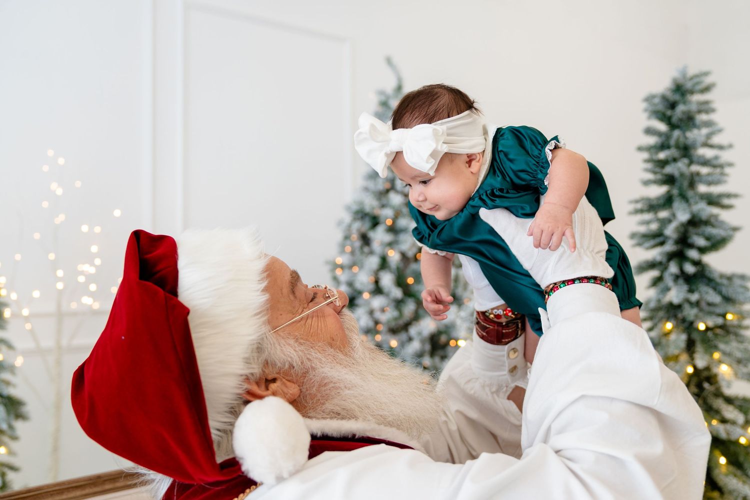 Santa Claus in red suit holds smiling baby in green dress up near decorated Christmas tree with twinkling lights.