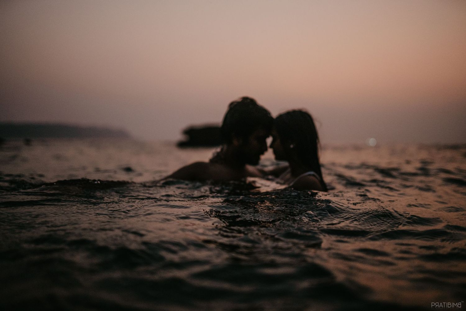 Romantic couple sharing an intimate moment in ocean water at sunset, silhouetted against a pink dusk sky.