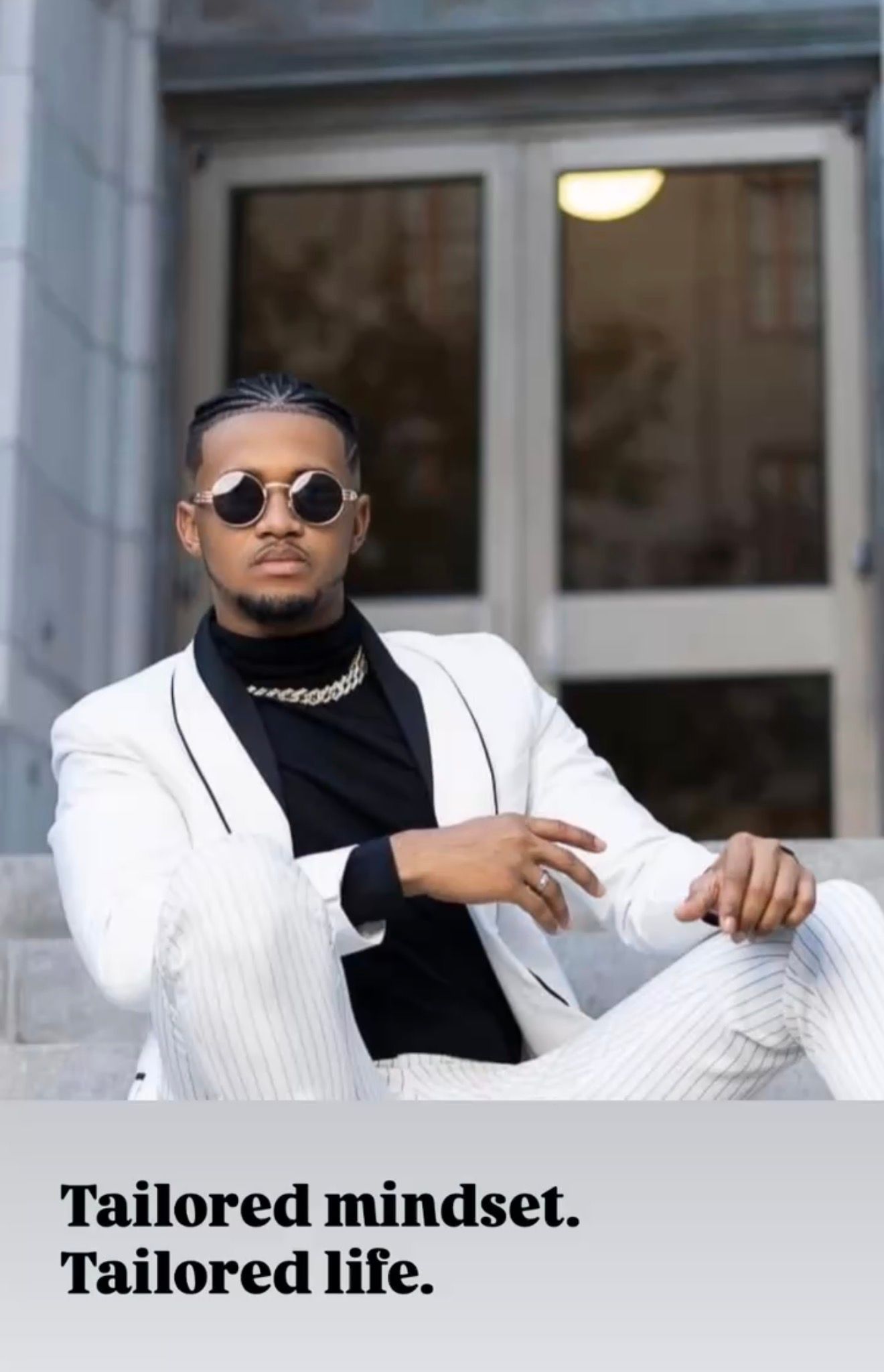 Stylish man in white pinstripe suit, black turtleneck, chain necklace, and round sunglasses sitting confidently on steps.